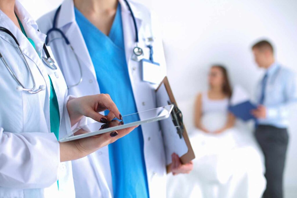 Healthcare professionals using a tablet, with a patient consultation in the background.